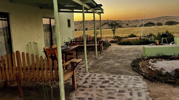 Un espacio de estar al aire libre cubierto, con suelo de piedra, muebles de madera y vistas a un campo al atardecer.