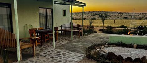 A covered outdoor patio with a stone floor, wooden furniture, and a view of a field at sunset.