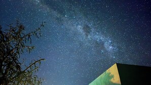 Cielo nocturno con estrellas y la Vía Láctea visibles sobre un edificio de techo inclinado.