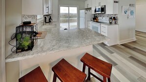 A modern kitchen with white cabinets, a marble countertop, and a view to the outside. There are two wooden stools and a small kitchen island.