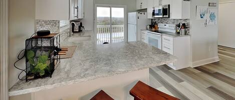 A modern kitchen with white cabinets, a marble countertop, and a view to the outside. There are two wooden stools and a small kitchen island.