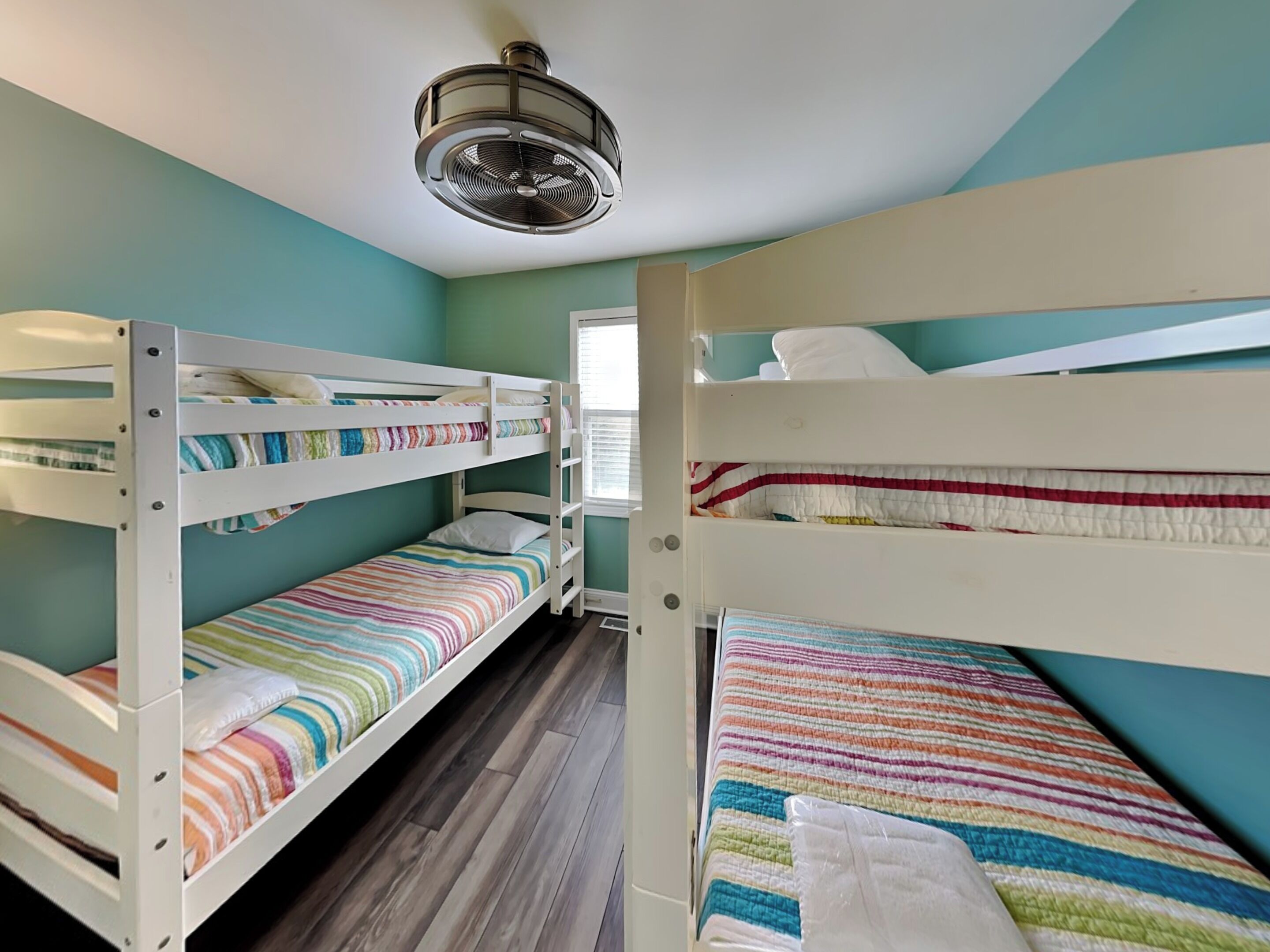 Une chambre avec un lit superposé, un ventilateur de plafond, un plancher en bois et des murs de couleur turquoise.