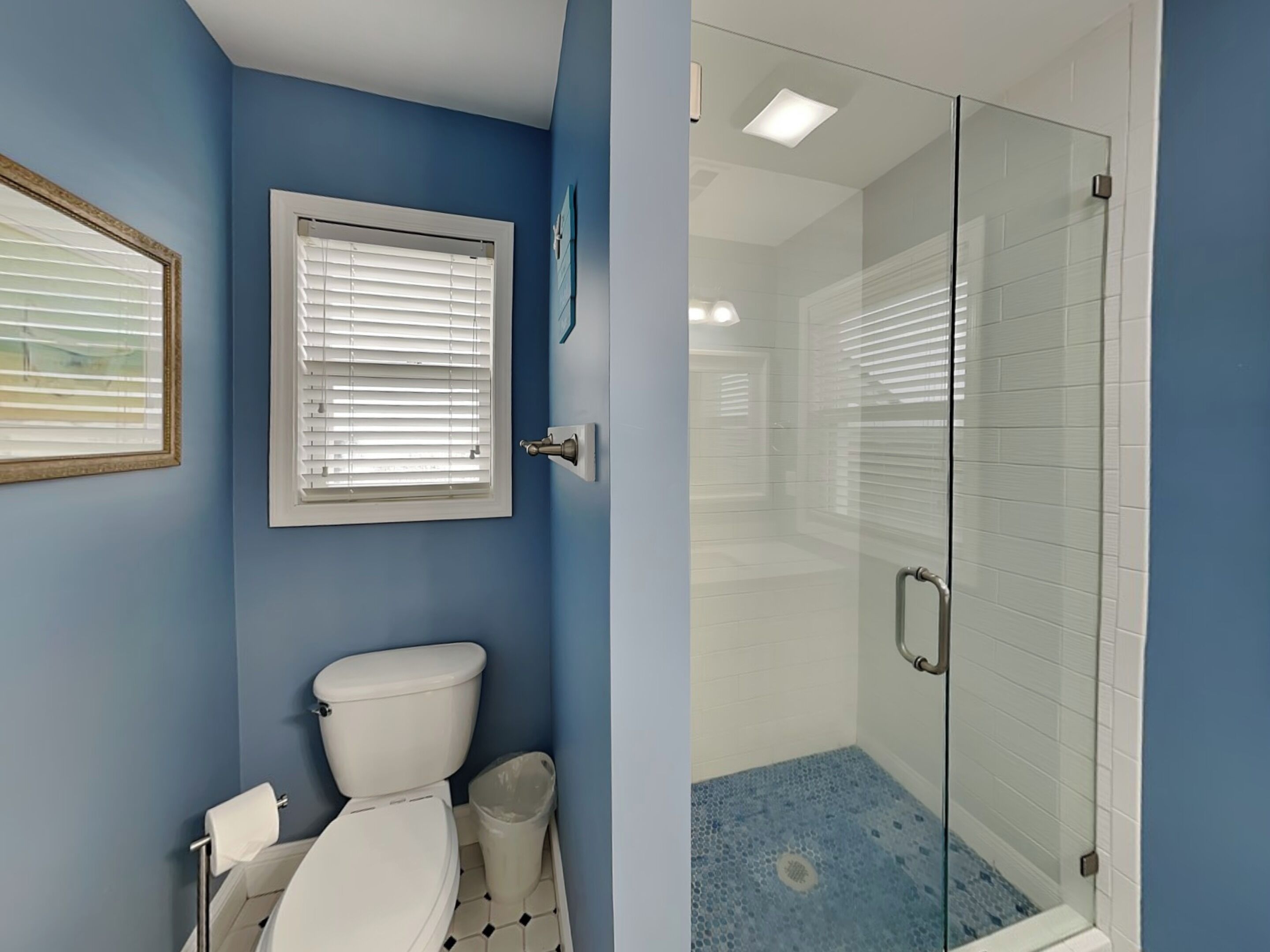A bathroom with a toilet, a window with blinds, and a glass shower enclosure.