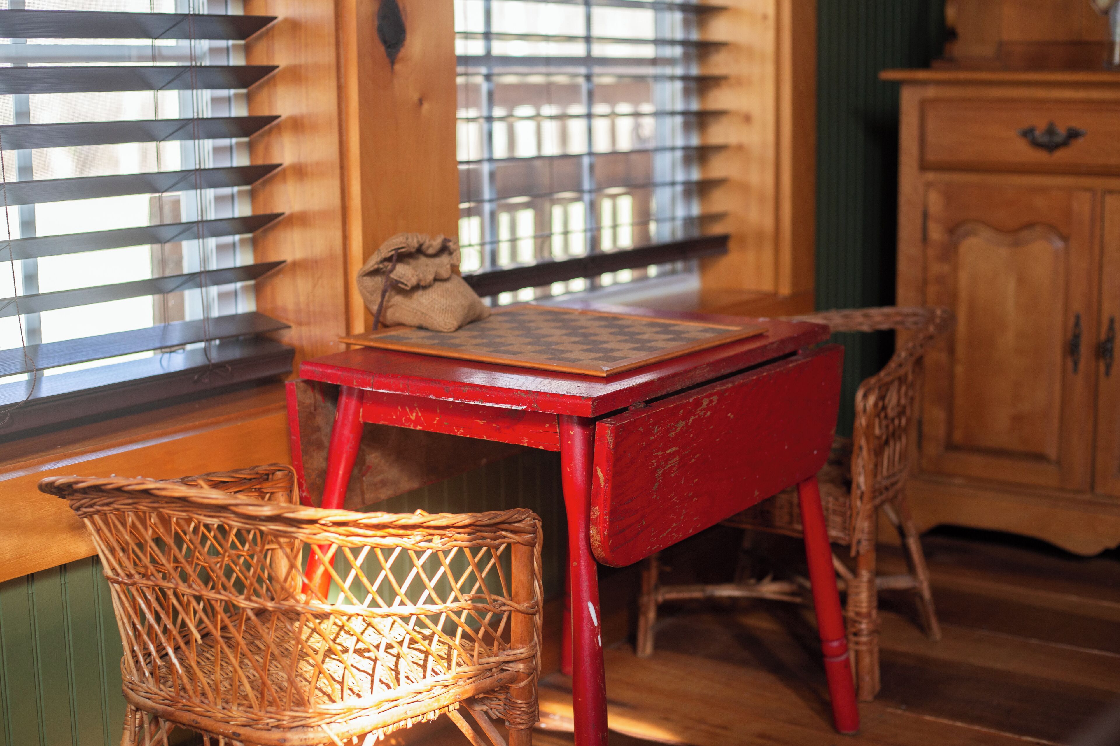 A wooden table with a woven basket and a small sack on top, a wicker chair, and a wooden cabinet in a room with wooden blinds and a window.