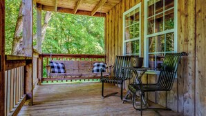 A wooden porch with a bench and chairs, large windows, and a view of trees.