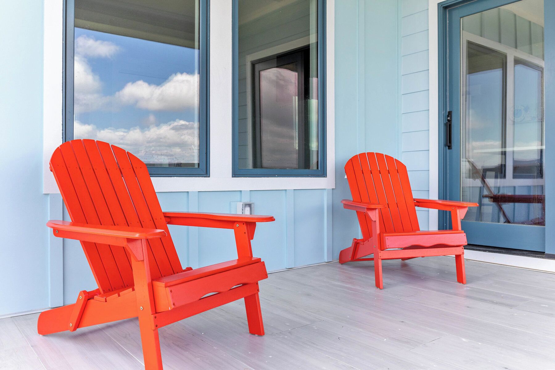 Dos sillas Adirondack rojas sobre una terraza de madera con una pared exterior azul claro y ventanas grandes.