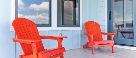 Dos sillas Adirondack rojas sobre una terraza de madera con una pared exterior azul claro y ventanas grandes.