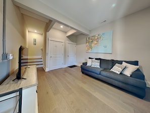 A spacious living room with a large blue sofa, wooden flooring, and a map on the wall. - Family Tides by Topsail Realty (Sneads Ferry)