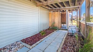 A covered patio with a wooden ceiling, a concrete pathway, and a small garden area with red mulch and plants. - In Good Sands by Topsail Realty (Sneads Ferry)