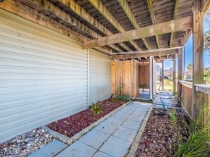 A covered patio with a wooden ceiling, a concrete pathway, and a small garden area with red mulch and plants. - In Good Sands by Topsail Realty (Sneads Ferry)