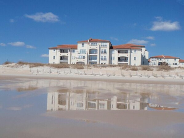 Une propriété située en bord de mer, dotée d’une piscine à eau calme et réfléchissante, de plusieurs balcons et d’une plage de sable en arrière-plan.
