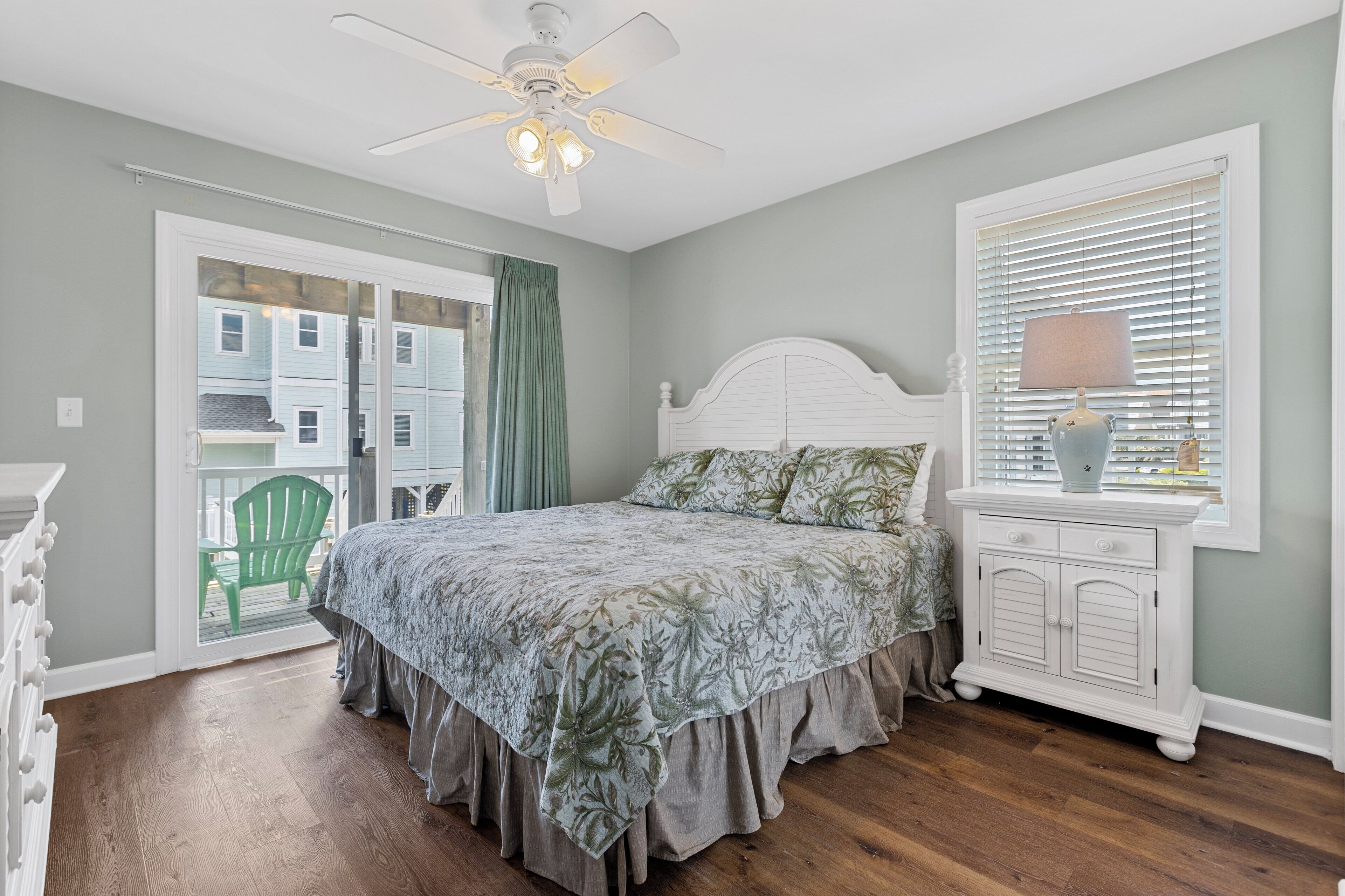 Une chambre avec un lit, une table de chevet, une fenêtre avec des stores, un ventilateur de plafond et une vue sur un fauteuil vert à l’extérieur.