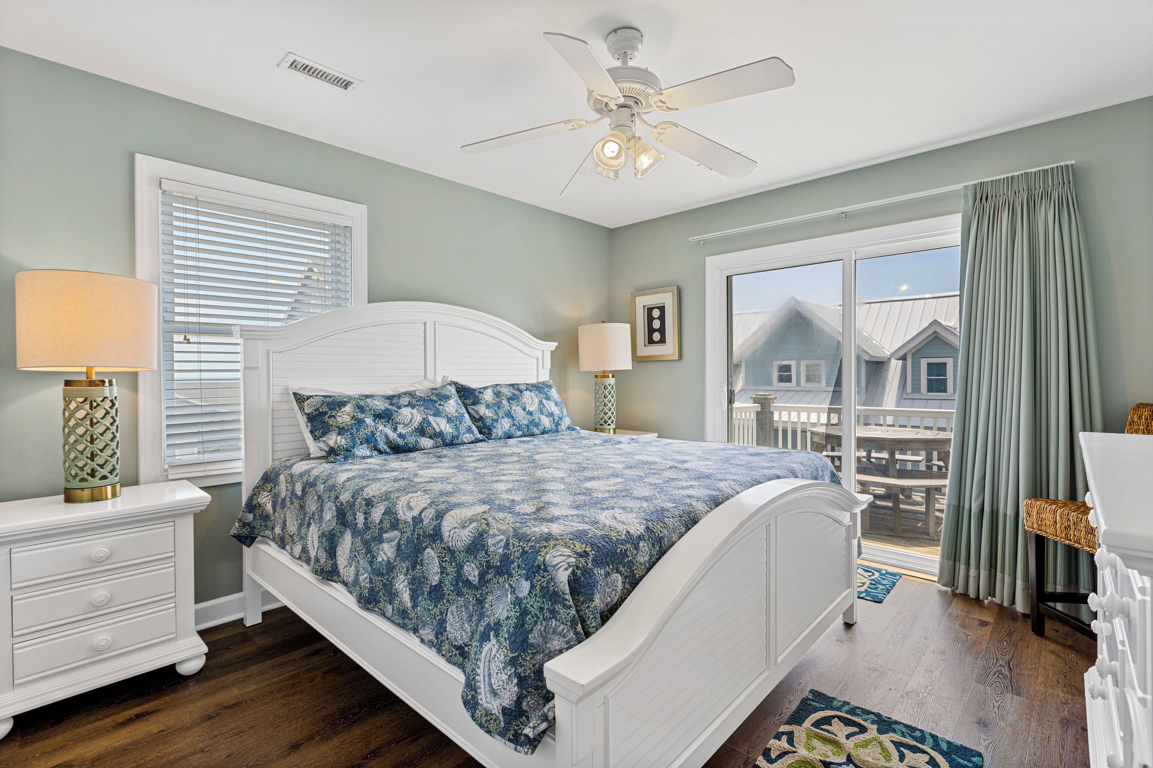 A bedroom with a white bed, a ceiling fan, a window with blinds, and a view of a balcony.