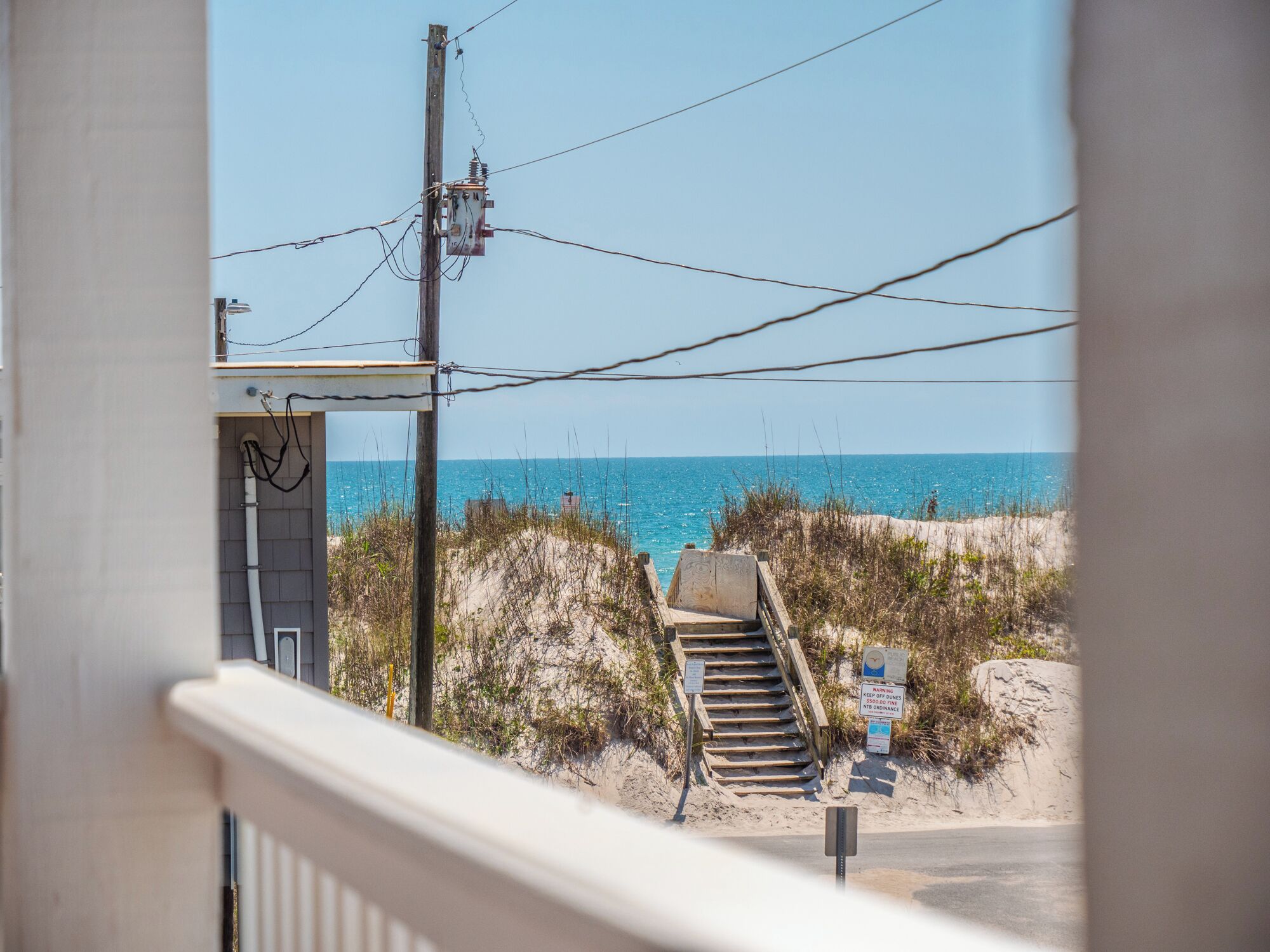 Uitzicht vanaf een balkon op een strand, met een trap die naar het strand leidt, een zandduin en een waarschuwingsbord over duinen.
