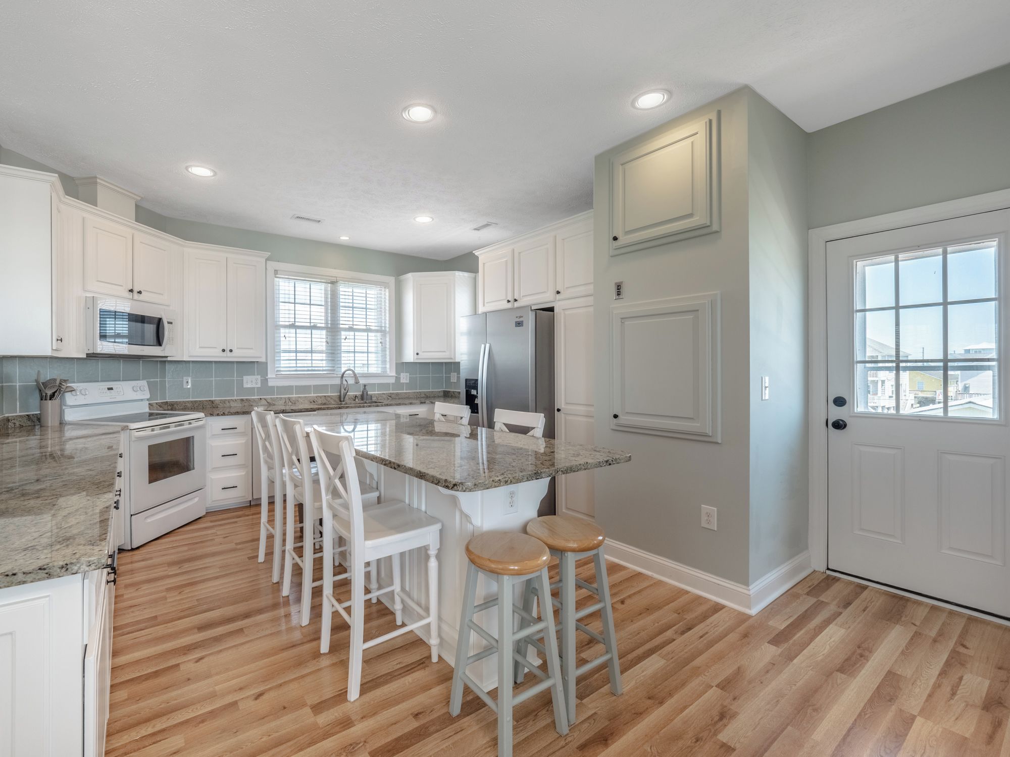 A modern kitchen with white cabinetry, a granite countertop, a stainless steel refrigerator, and a large window providing natural light.