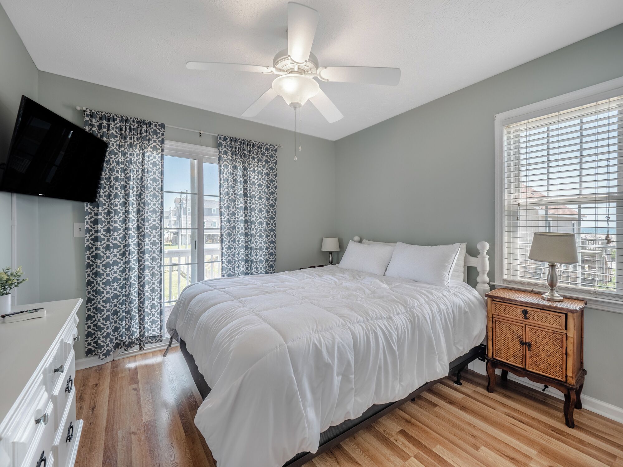 A bedroom with a bed, a ceiling fan, a TV mounted on the wall, a window with blinds, and a bedside table with a lamp.