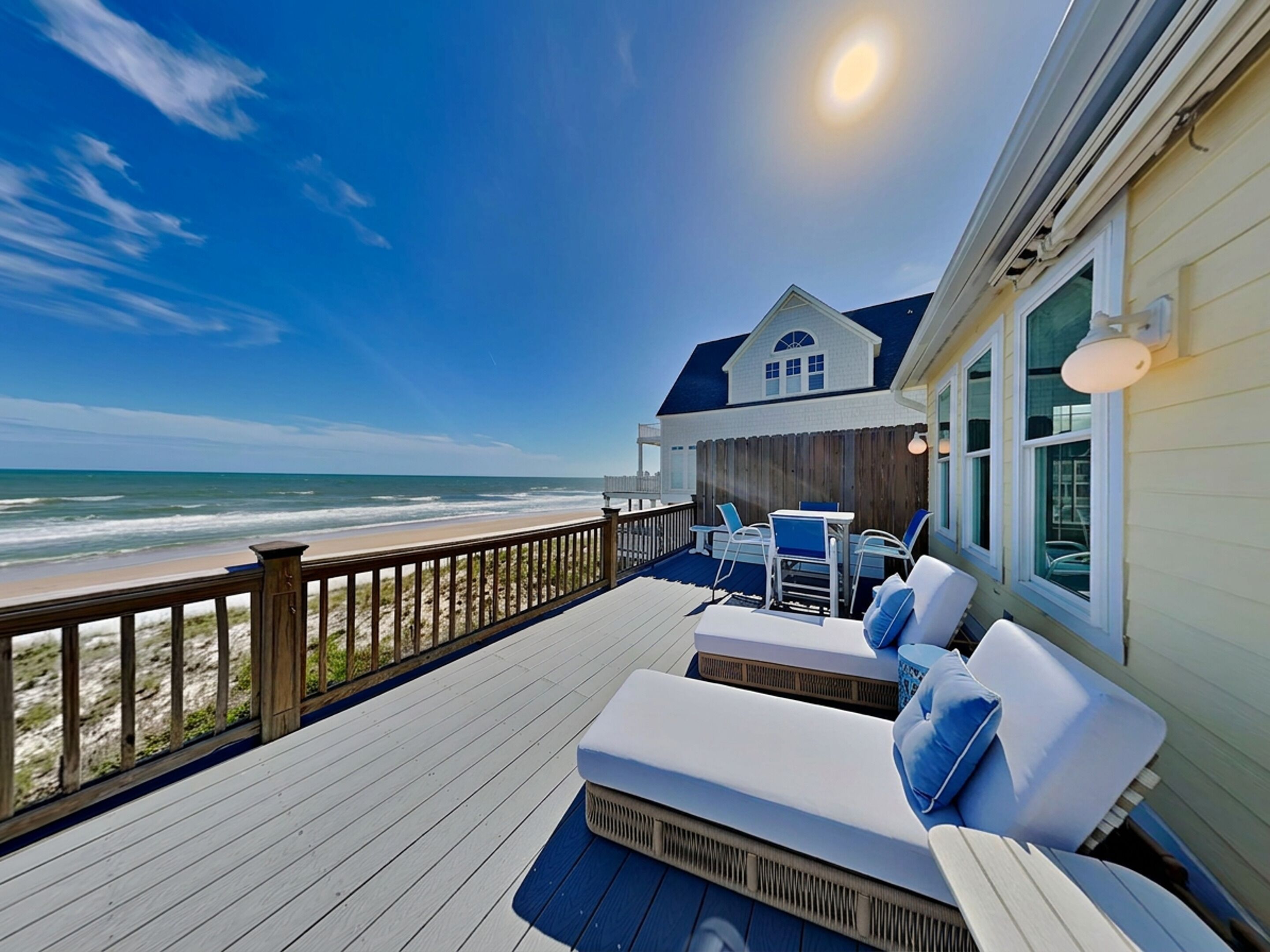A wooden deck with a view of the ocean, a white building with a blue roof, and outdoor furniture.