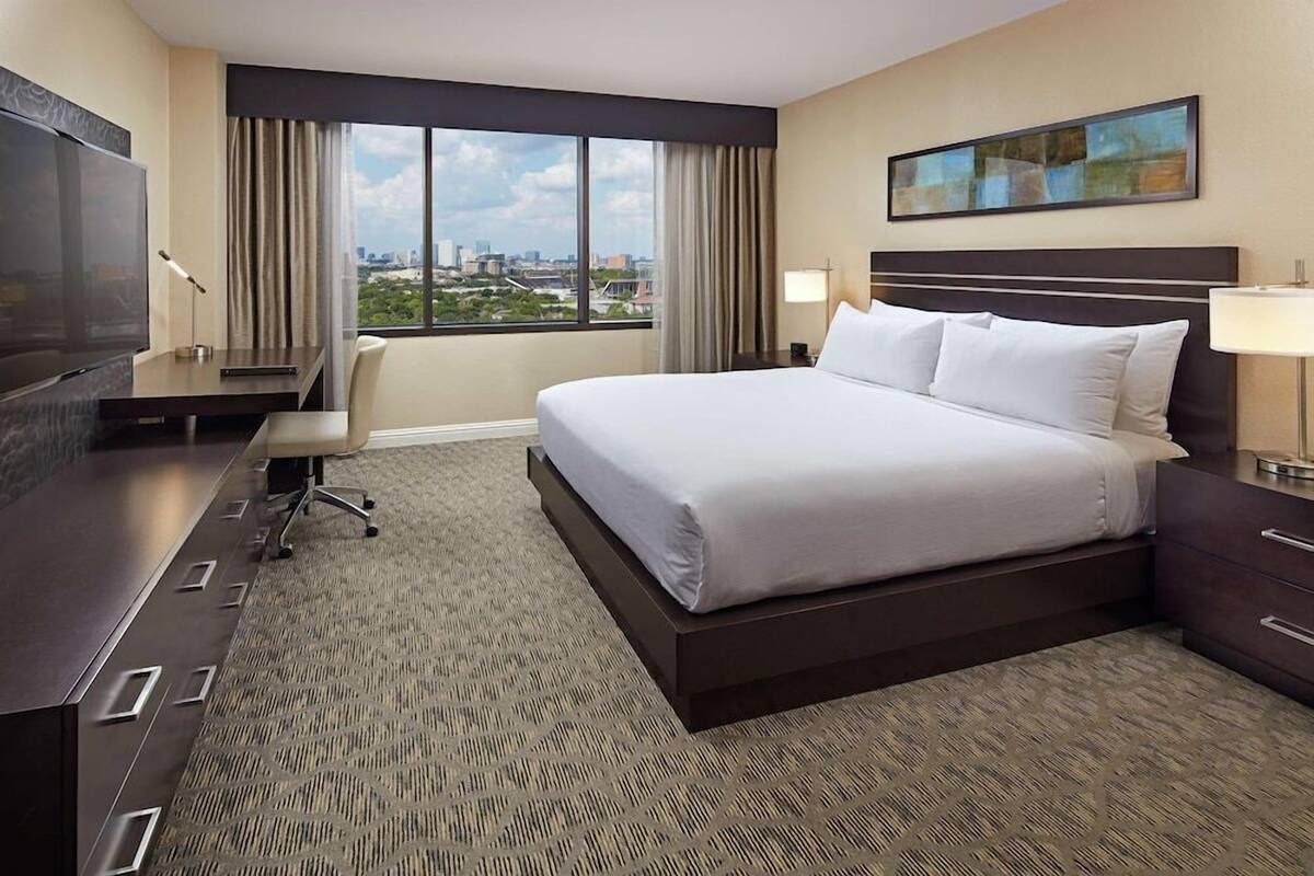 Ein Hotelzimmer mit einem großen Bett, einem Schreibtisch, einem Stuhl, einem Fernseher und Blick auf die Stadt.