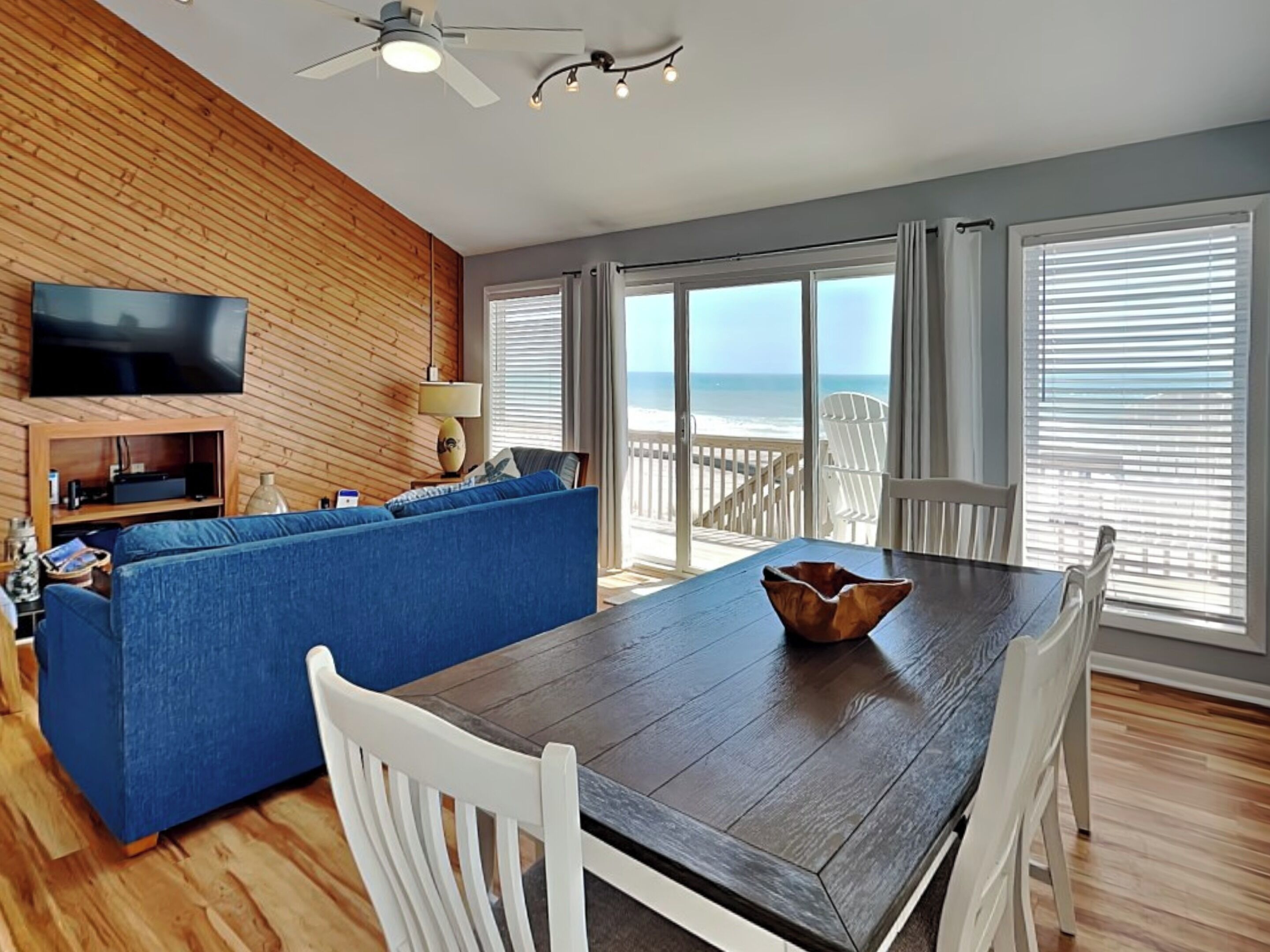 A modern living room with a wooden wall, a blue sofa, a wooden dining table with white chairs, and a TV mounted on the wall.