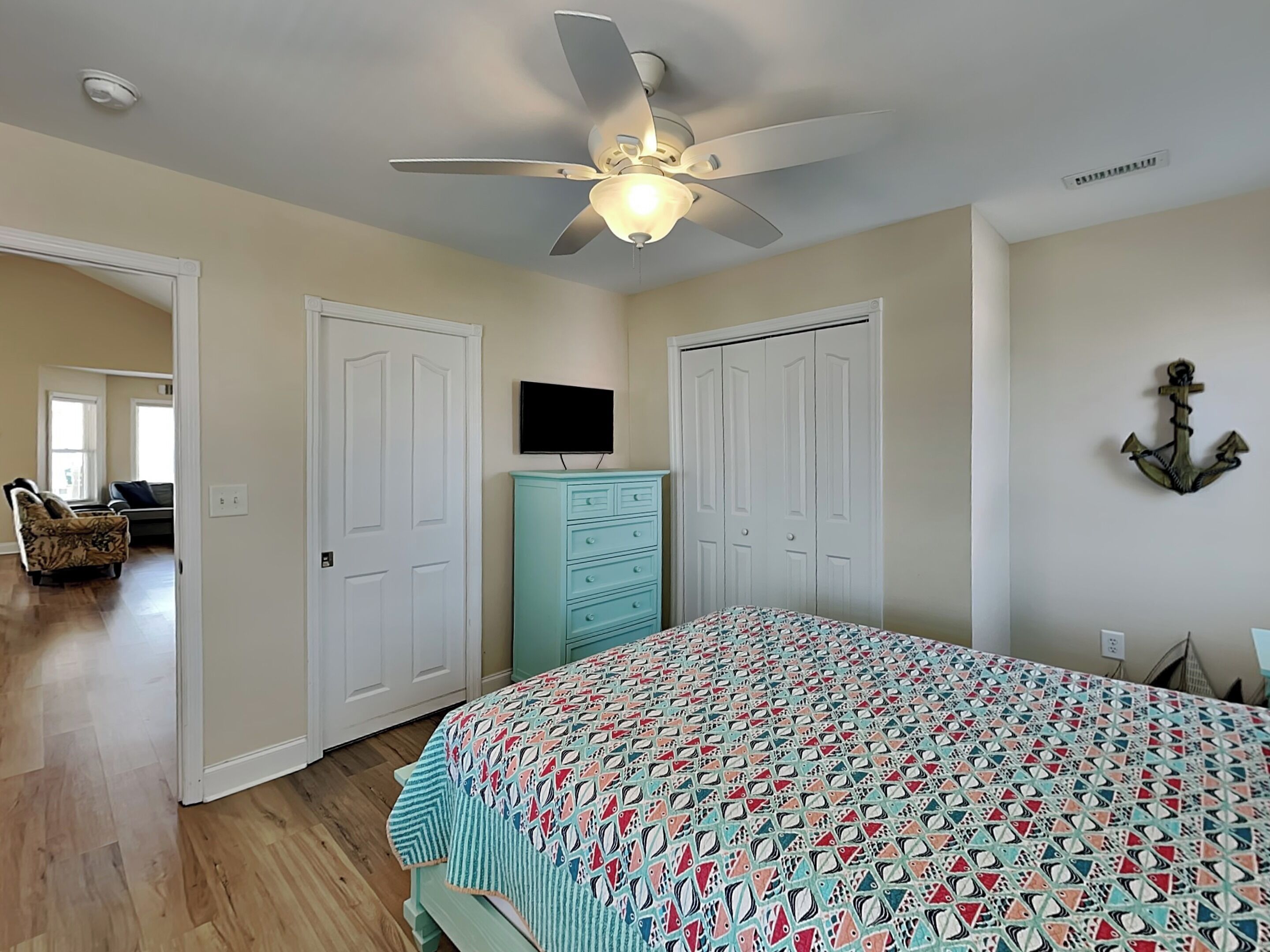 A bedroom with a ceiling fan, a bed with a patterned bedspread, a dresser, a television on the wall, and a closet.