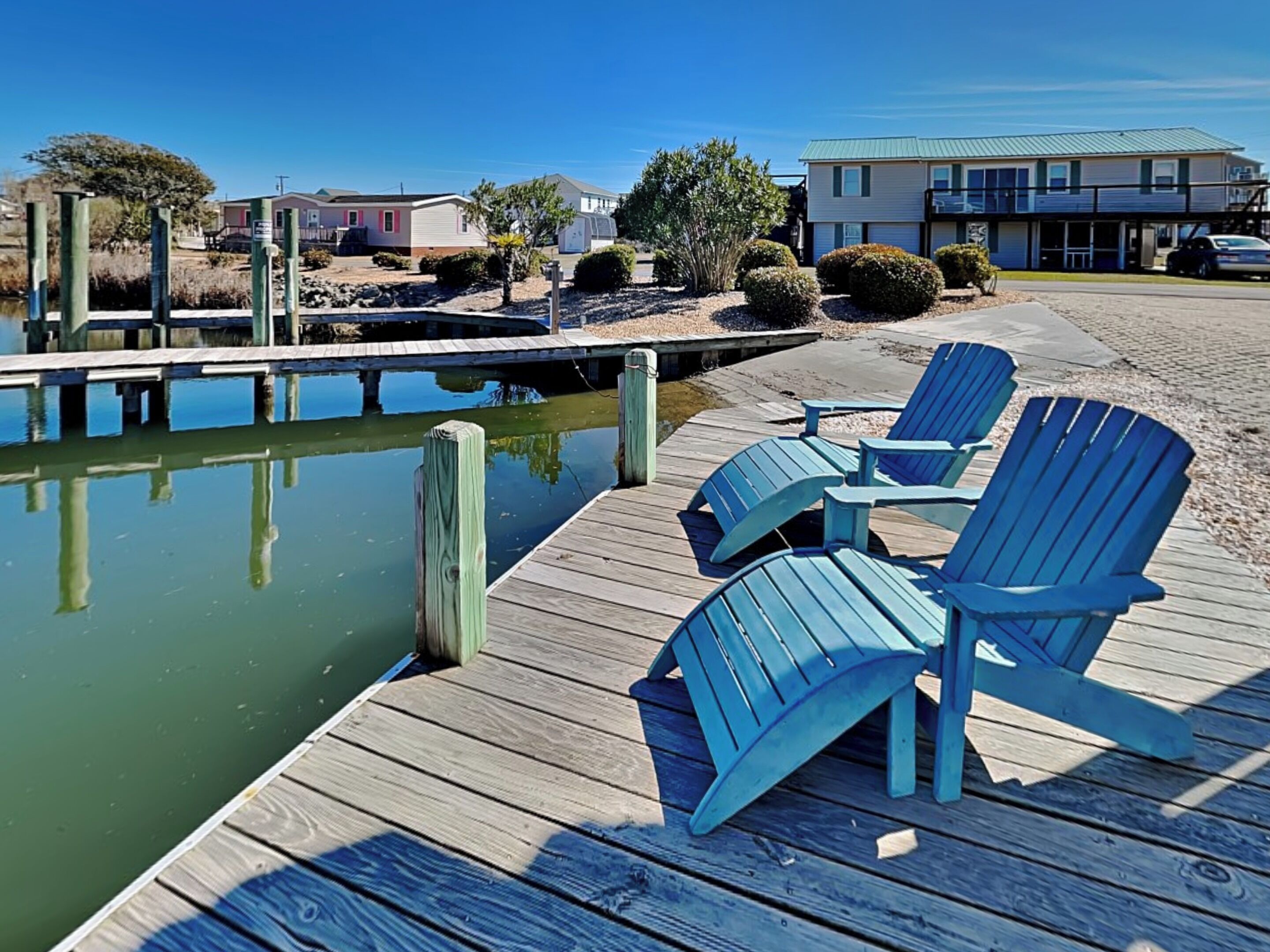 A wooden dock with blue Adirondack chairs, a clear view of the water, and a building in the background.