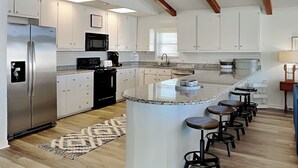 A modern kitchen with white cabinets, a stainless steel refrigerator, a black microwave, and a granite island with bar stools. - Bellcove by Topsail Realty (Holly Ridge)