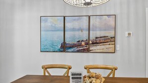 A dining area with a wooden table, two wooden chairs, a welcome sign, and a wall-mounted three-panel painting of a coastal scene. -   New Construction!!! Sleeps 7! Short drive to Sunset &Ocean Isle Beach / Golf!! (Sunset Beach)