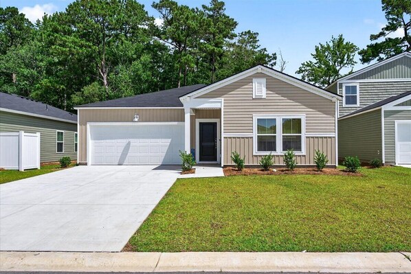 A single-story house with a garage, a front door, and a small porch with potted plants. -   New Construction!!! Sleeps 7! Short drive to Sunset &Ocean Isle Beach / Golf!! (Sunset Beach)