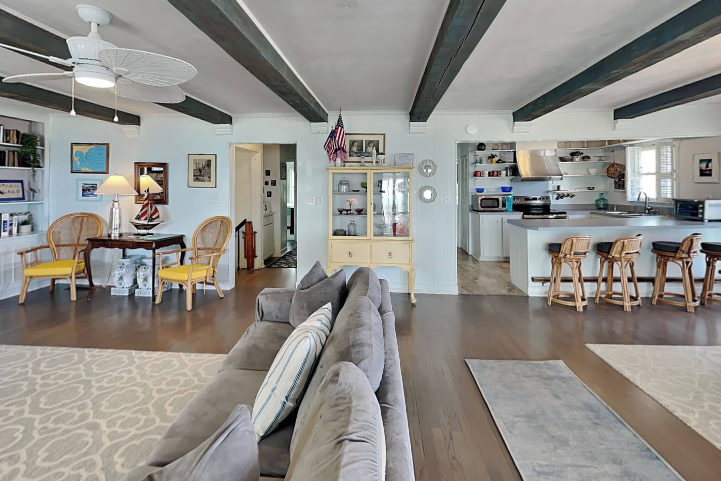 A spacious living room with a gray sofa, wooden chairs, and a dining area with a table and chairs. A kitchen with a large island and bar stools is visible in the background.