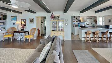 A spacious living room with a gray sofa, wooden chairs, and a dining area with a table and chairs. A kitchen with a large island and bar stools is visible in the background.