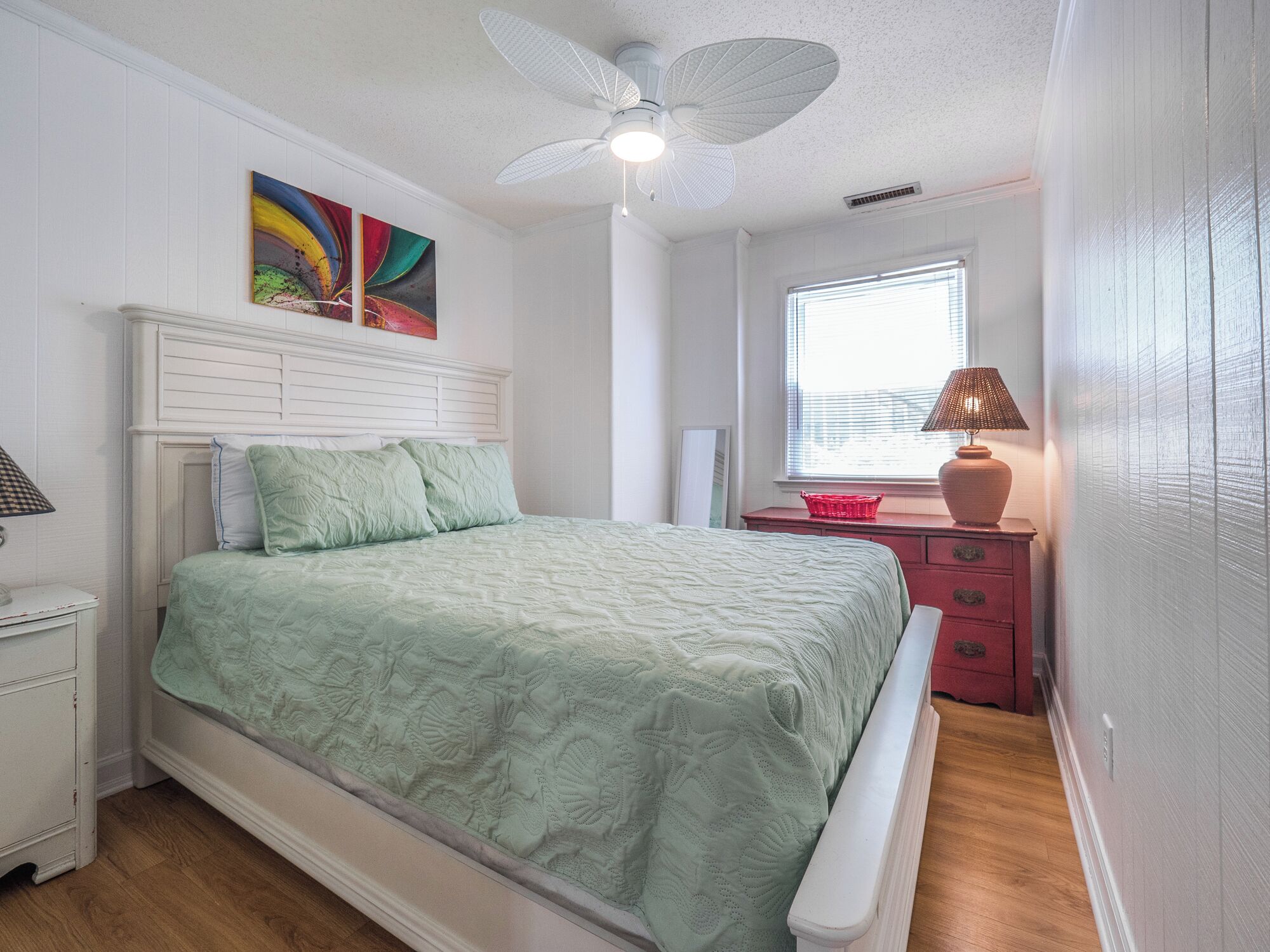 Un dormitorio con cama, mesita de noche, lámpara, ventana con persianas y ventilador de techo.