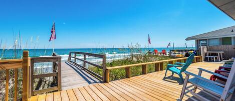 A wooden deck with a view of the ocean, featuring a beach house, beach chairs, and American flags.