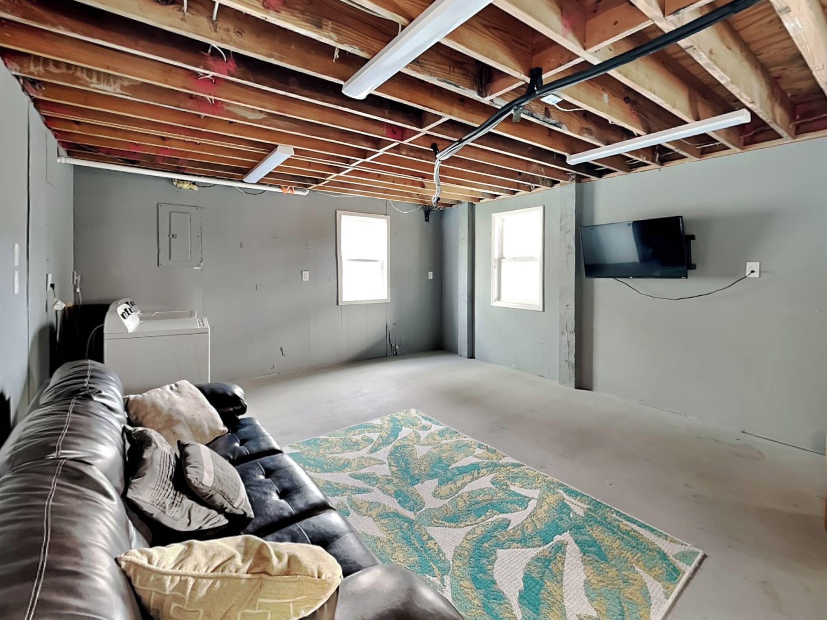 A spacious room with a high ceiling, exposed wooden beams, a black leather couch, a patterned rug, and a mounted TV.