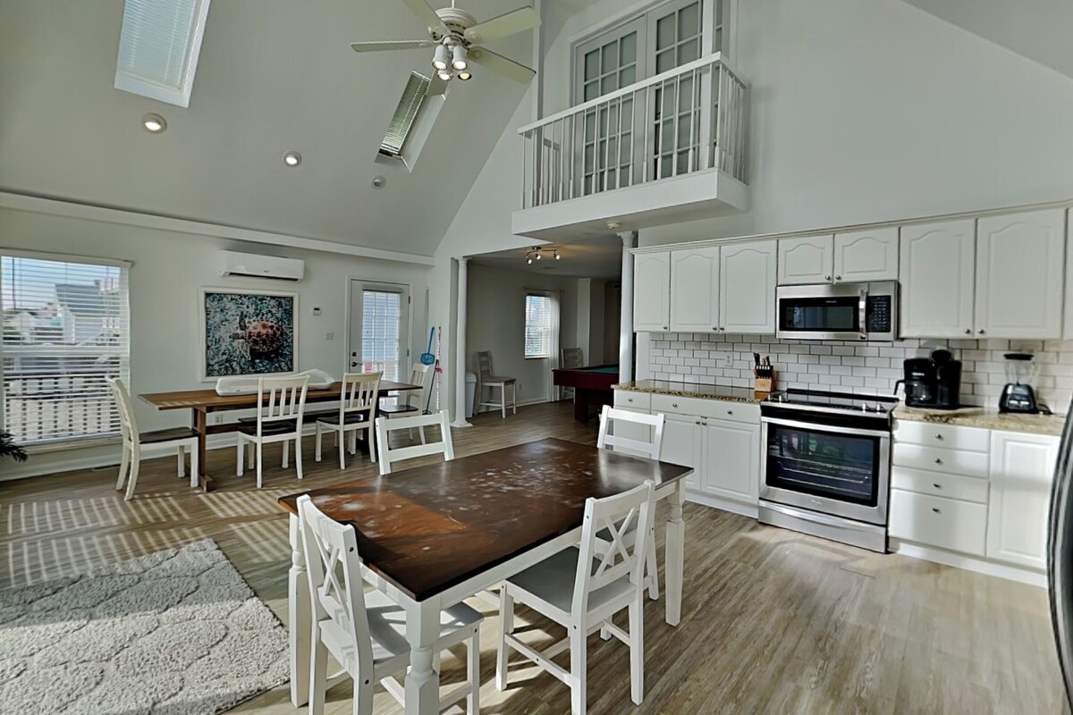 A spacious kitchen with white cabinets, a dining area with a wooden table and white chairs, and a pool table in the background.