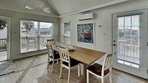 A dining area with a wooden table and white chairs, large windows with blinds, an air conditioning unit, and a door with a view.