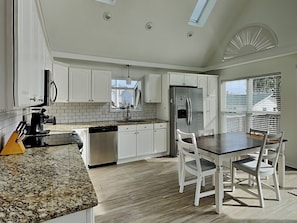 A modern kitchen with white cabinetry, a granite countertop, a stainless steel refrigerator, and a dining area with white chairs.