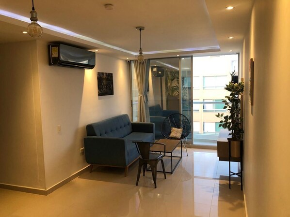 Un salon moderne avec un canapé, une table basse et un fauteuil. Une grande fenêtre avec des rideaux, une plante et un appareil de climatisation fixé au mur complètent la pièce.