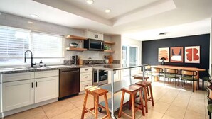 A modern kitchen with white cabinets, a stainless steel dishwasher, a microwave, and a stove. There are wooden stools and a dining table with chairs. - 5BR Fun Haven: Pool, Mini Golf, Movie, Gameroom (Riverside)