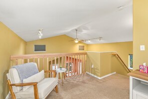 A room with a sloped ceiling, a wooden chair, a small table, a ceiling fan, and a staircase leading to another level.