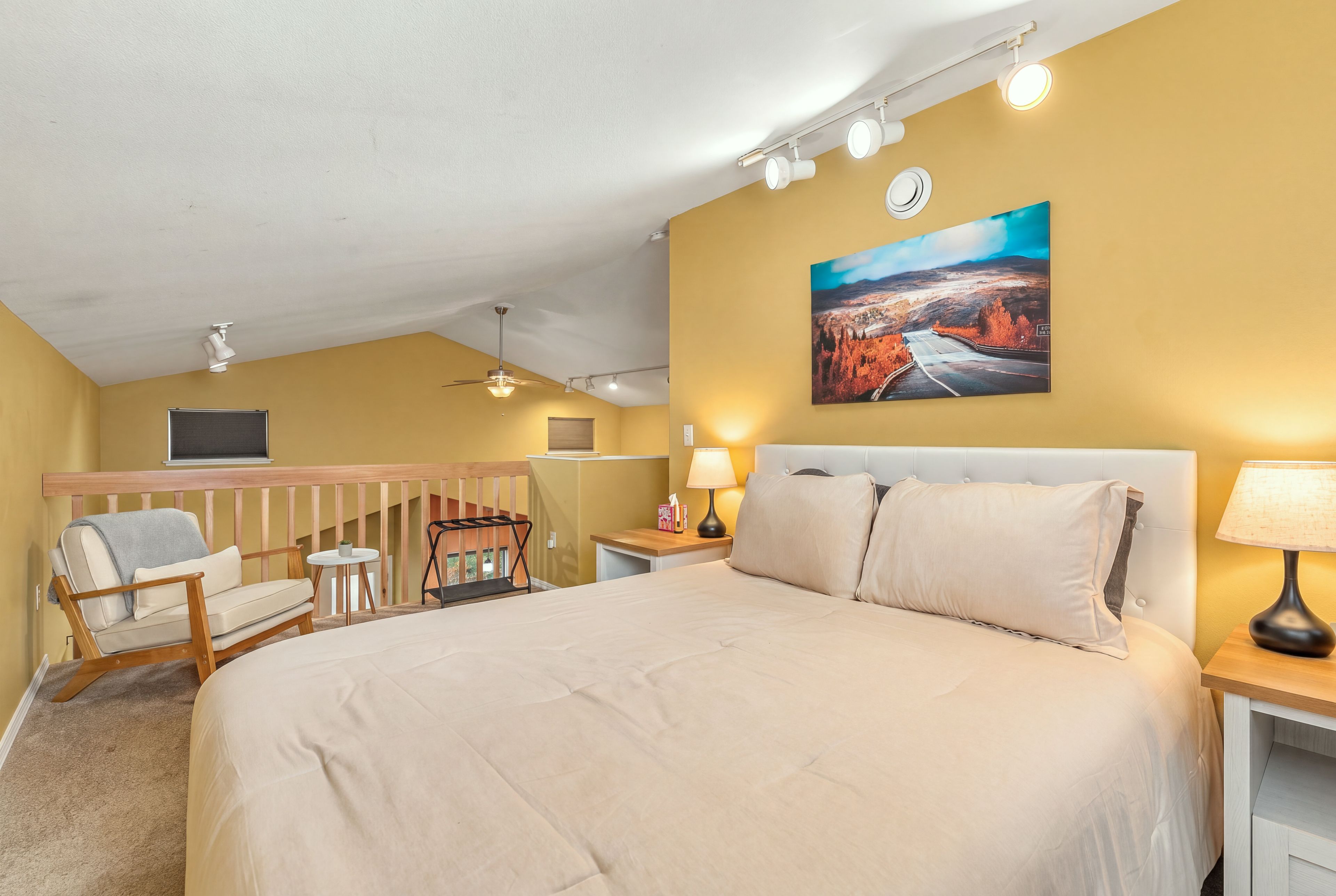 Une chambre d’hôtel avec un grand lit, une chaise, un bureau, un tableau au mur et un ventilateur de plafond.