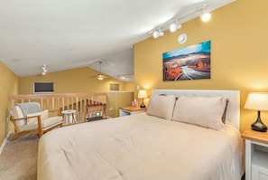 Une chambre d’hôtel avec un grand lit, une chaise, un bureau, un tableau au mur et un ventilateur de plafond.