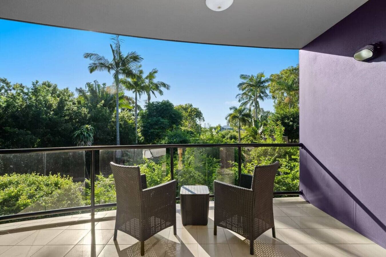 Un balcon agrémenté de fauteuils et d’une table en osier, donnant sur un jardin luxuriant planté de palmiers.
