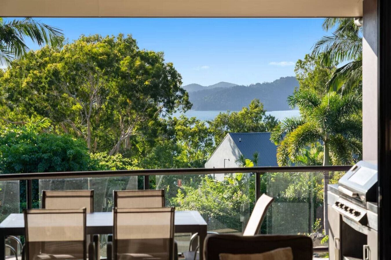Ein Balkon mit Blick auf ein Gewässer, Bäume und ein ferneres Gebirge. Auf dem Balkon befinden sich Stühle und ein Grill.