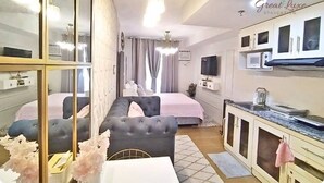 A modern hotel room with a bed, a sitting area with a chair and table, a kitchenette, and a large mirror.