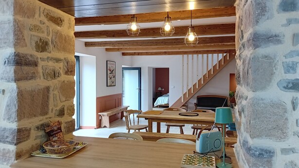 Un comedor acogedor con una mesa de madera, sillas y una pared de piedra. Se observan lámparas colgantes y una escalera al fondo.
