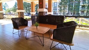 Un área de estar al aire libre con mobiliario de mimbre y una mesa de madera, con vistas a un edificio residencial y coches estacionados al fondo.