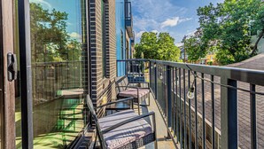 Un balcon doté d’une barrière en verre, d’un espace salon extérieur et offrant une vue sur une rue bordée d’arbres.