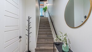 Un couloir avec un escalier en bois, une porte blanche, un porte-manteau, un miroir et un vase rempli de fleurs.