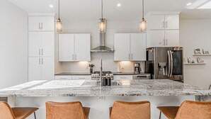 Une cuisine moderne avec des meubles blancs, un îlot central doté de tabourets, des appareils en acier inoxydable et un éclairage suspendu.
