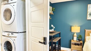 A laundry room with stacked washers and dryers, a wooden table with chairs, a nightstand with a lamp, and a bed with a headboard. - Private Studio/1 Min-Hospital/Self Check-In/75" TV (Williams Lake)